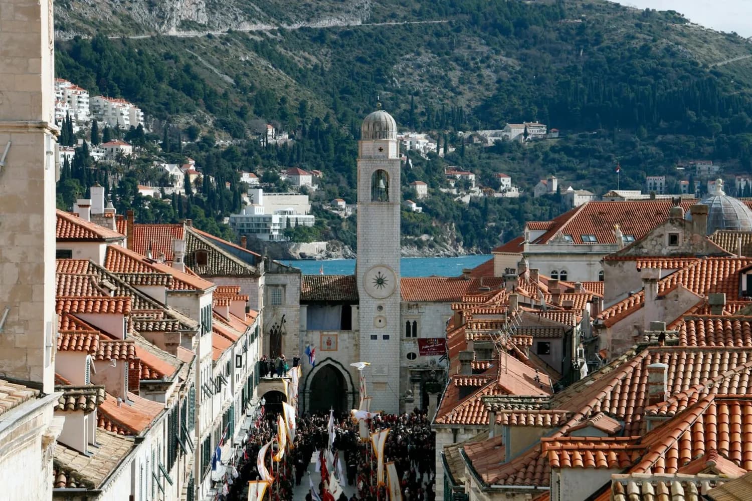 Blick auf die Altstadt von Dubrovnik mit roten Dächern, Uhrturm und belebter Hauptstraße Richtung Meer und bewaldeten Hügeln im Hintergrund
