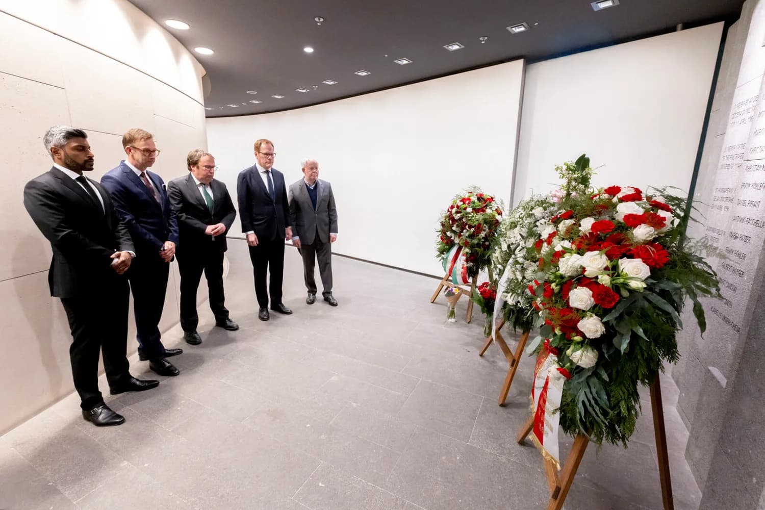 Fünf Männer in Anzügen stehen schweigend vor Kranzarrangements mit roten und weißen Blumen in einer Gedenkstätte - Landesminister Oliver Krischer (Mitte), Oberbürgermeister Dr. Stephan Keller (zweiter von rechts), Dr. Rolf Pohlig (rechts, stellvertretender Vorsitzender des Aufsichtsrats des Flughafens Düsseldorf) sowie Lars Redeligx (zweiter von links, Vorsitzender der Geschäftsführung) und Pradeep Pinakatt (links, kaufmännischer Geschäftsführer), anlässlich des 30. Jahrestages des Brandunglücks vom 11. April 1996 im Gedenkraum des Airports. Foto: Flughafen Düsseldorf / Andreas Wiese
