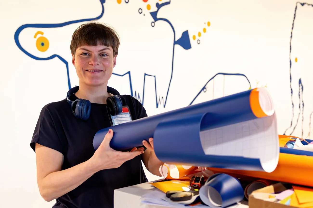 Künstlerin Anne Bracht hält blau-orangenes Papierrolle vor Wandgrafik, Bastelmaterial und Kopfhörer sichtbar