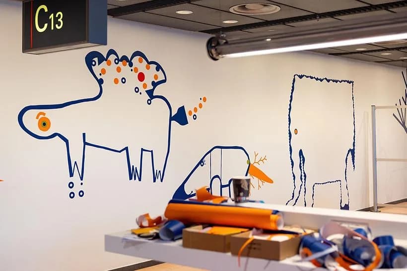 Wartebereich am Gate C13 mit bunten Wandillustrationen in blau und orange, stilisierte Tiermotive und Sitzbereich mit verstreuten Bastelmaterialien auf einem Tisch