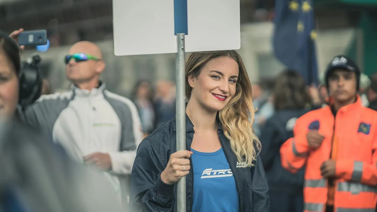 Junge Frau lächelt auf Streik‑Demonstration und hält Schild, Menschenmenge im Hintergrund mit Warnwesten und Fahne