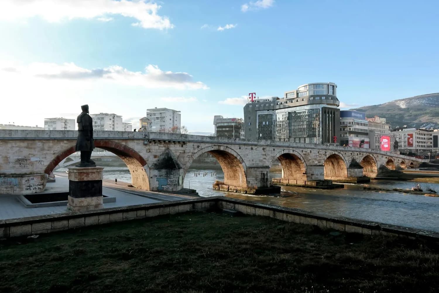 Steinbrücke über den Fluss Vardar in Skopje mit Bogengängen, Uferpromenade, Statue im Vordergrund und moderner Stadtbebauung im Hintergrund