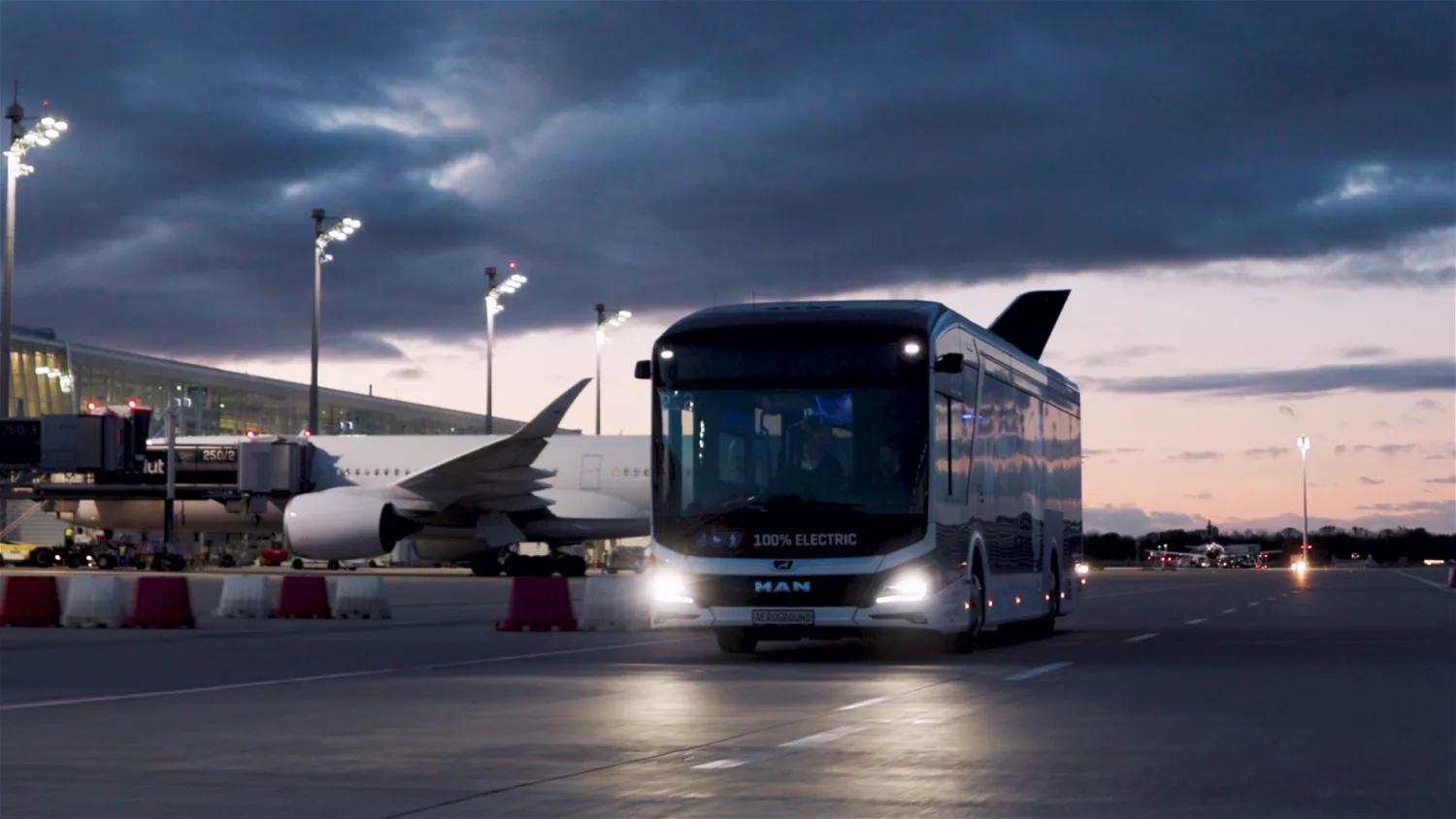 Elektro-Flughafenbus LionsCity e Aeroground vor Parkposition eines Flugzeugs bei Dämmerung, Scheinwerfer an und Passagierterminal im Hintergrund