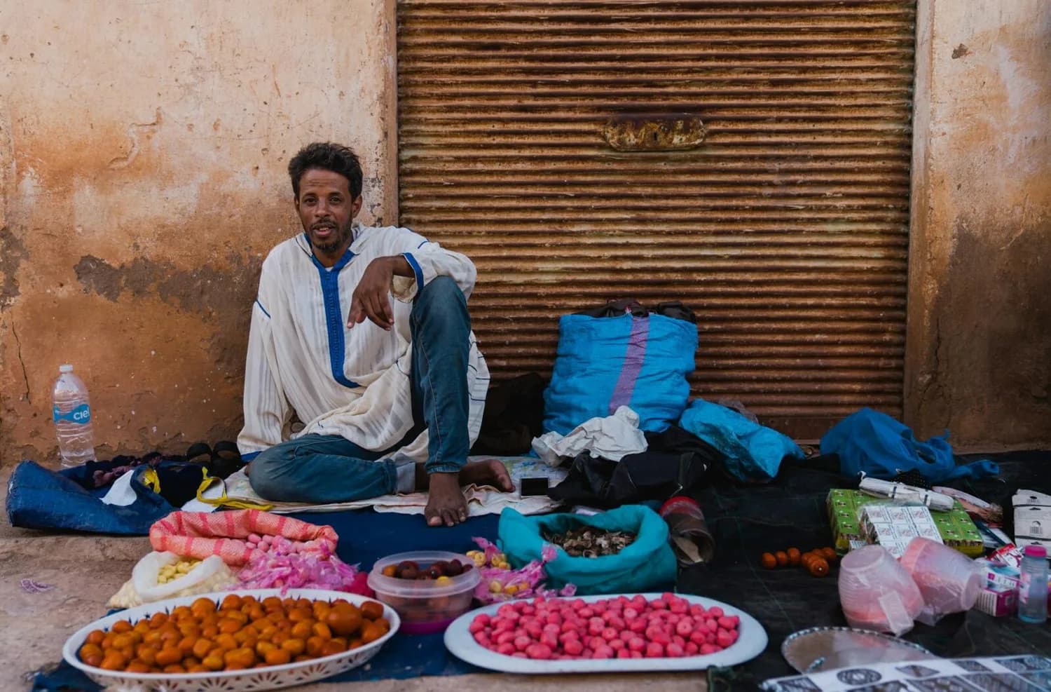 Marrakesch Straßenhändler sitzt vor geschlossener Metalltür und verkauft bunte Trockenfrüchte und Süßigkeiten auf ausgestellten Tabletts in Medina von Marokko