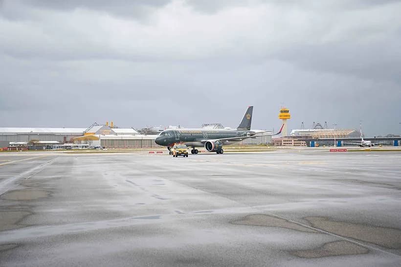 Royal Jordanian Passagierflugzeug in dunkler Sonderbemalung auf Startbahn bei Erstflug, Flughafen-Rollfeld mit Tower und Bodendienstfahrzeug bei bewölktem Wetter