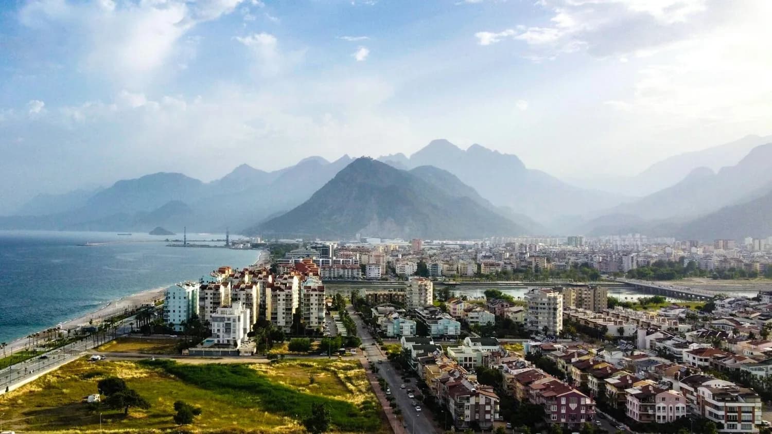 Blick auf Antalya Küste mit Wohngebäuden, Strandpromenade und Bergen im Hintergrund, Stadtszene mit Hafen und grünen Flächen