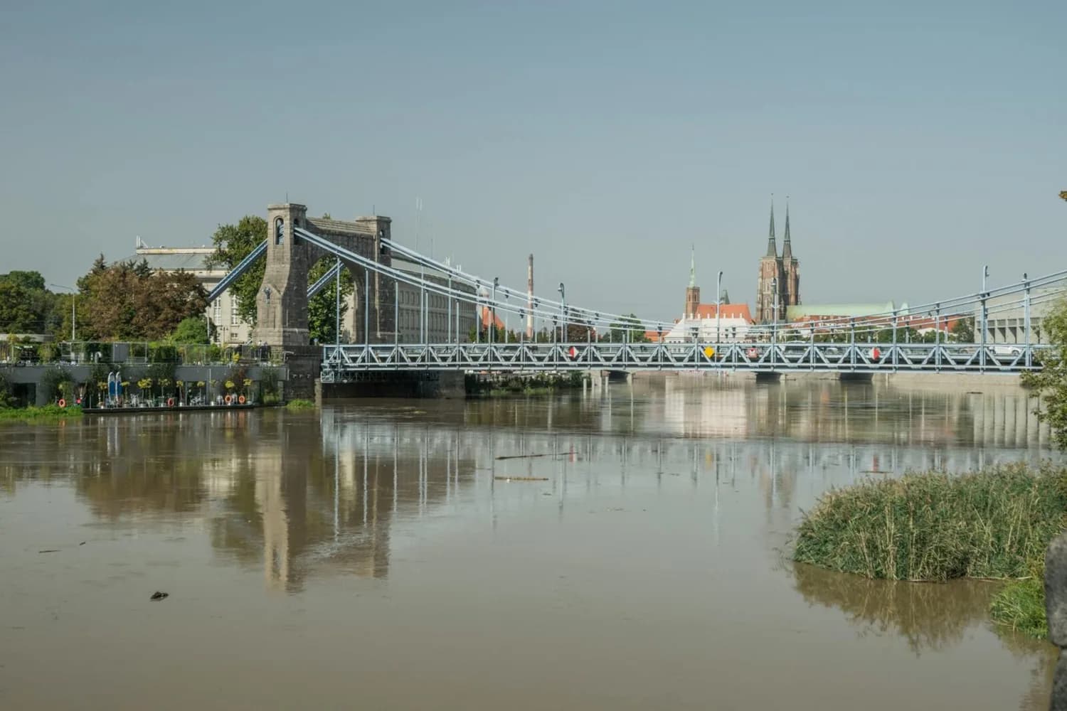Hängebrücke über ruhigen Fluss mit Uferpromenade und Stadtsilhouette von Breslau im Hintergrund, Blick auf Kirchtürme und Park-Sleep-Fly-Parkplatz nahe Dortmund-Flugverbindung