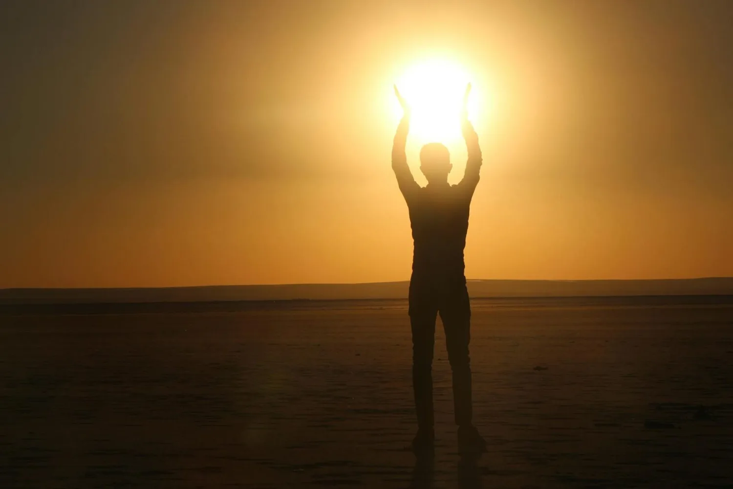Silhouette einer stehenden Person, die bei Sonnenuntergang die Hände zur Sonne hebt am Strand/weiter Küste, warmes Urlaubslicht