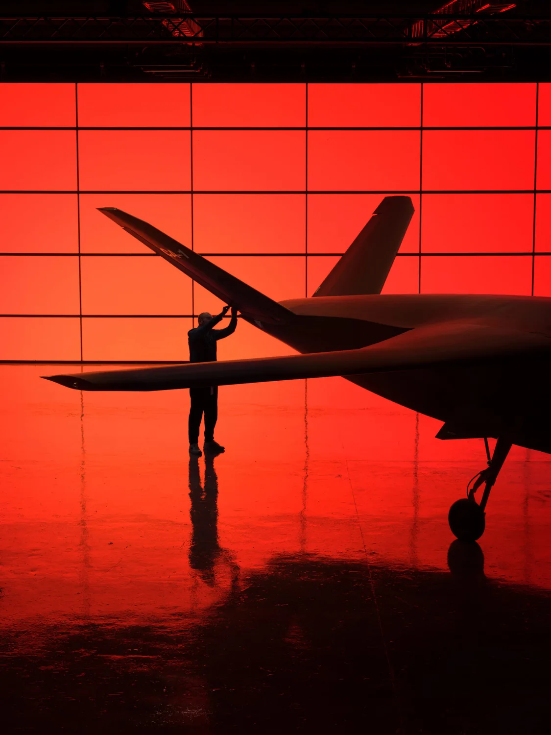 Person prüft Flugzeugflügel in Hangar vor leuchtend roter Wand, silhouette eines Flugzeugs und Reflexion auf glänzendem Boden