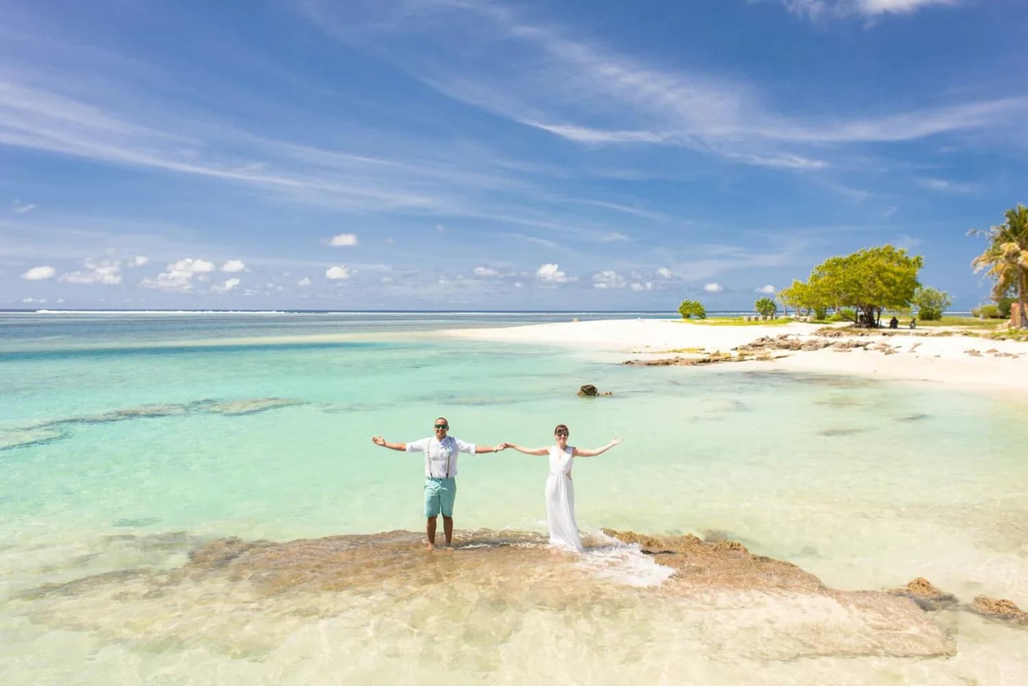 Braut und Bräutigam halten Händchen auf Felsen im flachen türkisfarbenen Wasser vor weißem Sandstrand auf Mauritius, romantische Strandhochzeit, Eurowings Marriage Package Hinweis