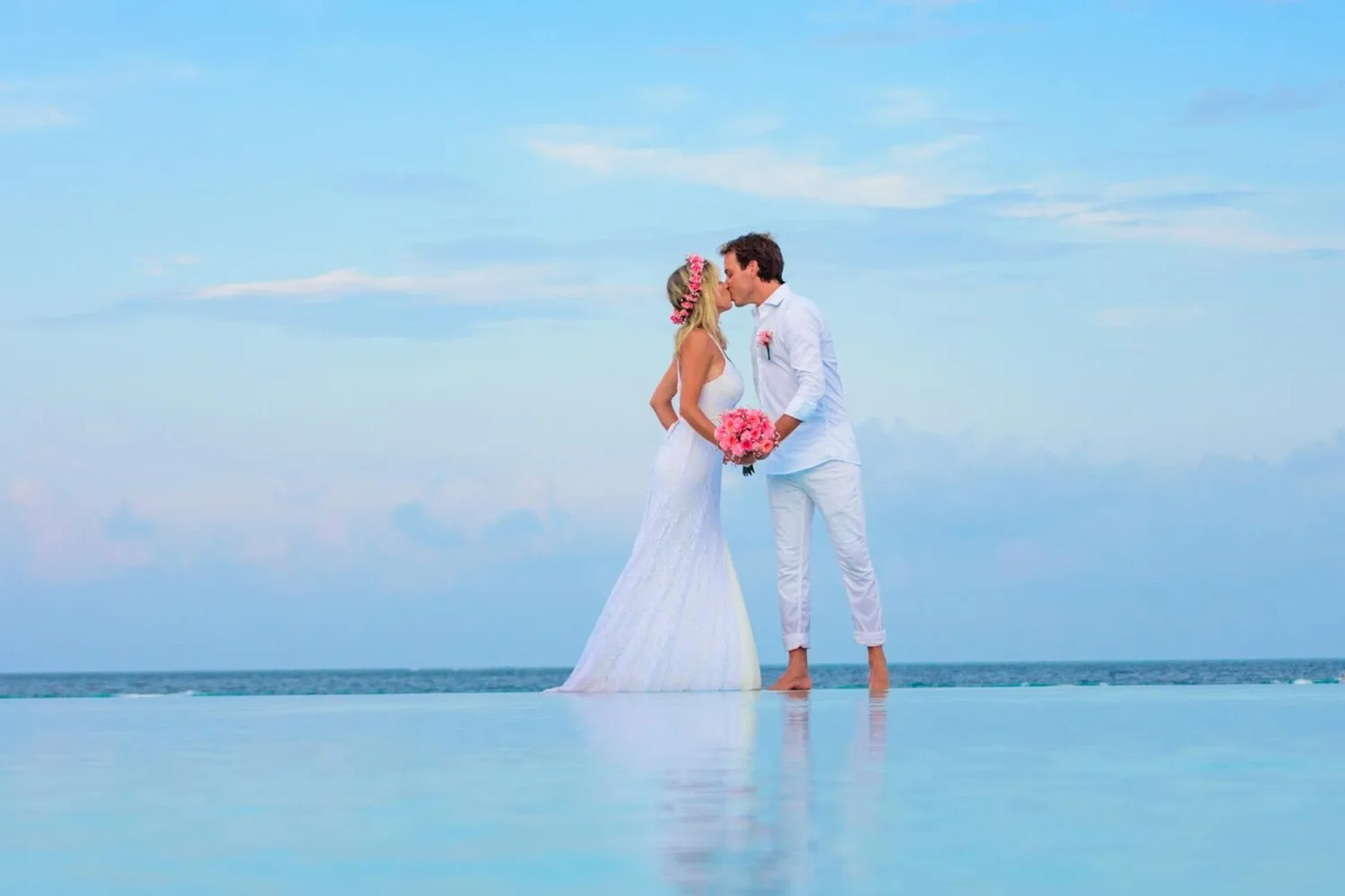 Brautpaar in weißer Kleidung küsst sich am Strand bei Meerblick, Braut mit Blumenkranz und rosa Blumenstrauß – Heiraten Marriage Package Eurowings