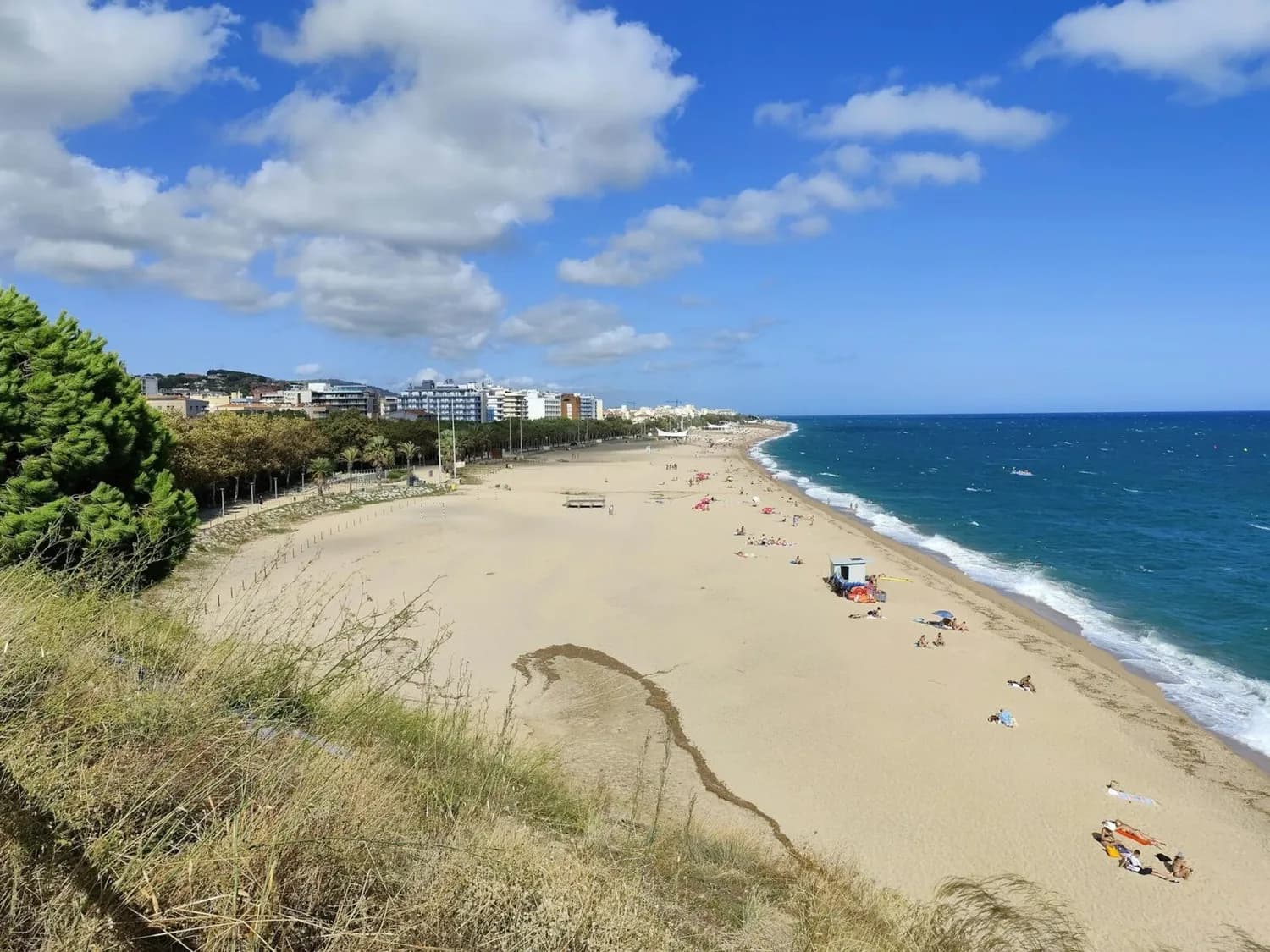 Sandstrand von Calella in Katalonien mit Badenden, blauem Mittelmeer, Strandpromenade und Stadtgebäuden im Hintergrund