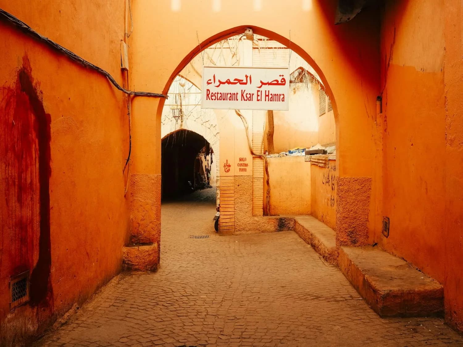 Eingangsbogen zur Gasse mit Schild Restaurant Ksar El Hamra, orangefarbene Mauern und gepflasterter Fußweg in Marrakesch