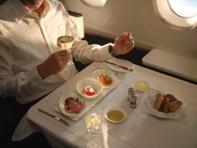 Mann in Business-Class neben Flugzeugfenster genießt Glas Weißwein und mehrgängige Gourmet-Vorspeisen mit Brot und Olivenöl auf Tisch mit weißer Tischdecke