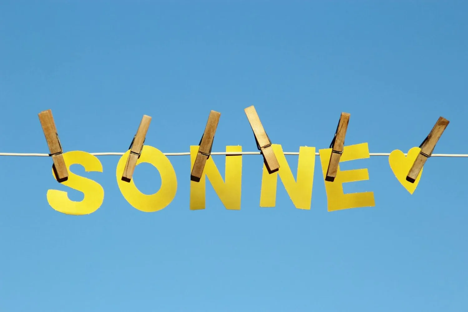 Gelbe Papierbuchstaben SONNE und Herz an Wäscheleine mit Wäscheklammern vor blauem Himmel – Symbol für Sonne, Osterferien, Flüge ab Nürnberg