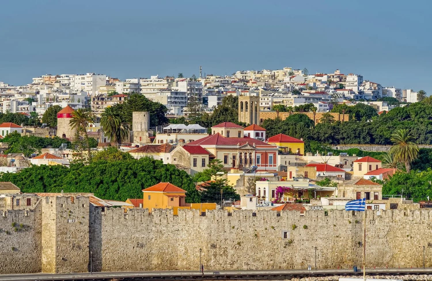 Blick auf die mittelalterliche Stadtmauer und bunte Häuser von Rhodos, Griechenland mit Palmen und griechischer Flagge, beliebtes Flugreiseziel ab Bremen