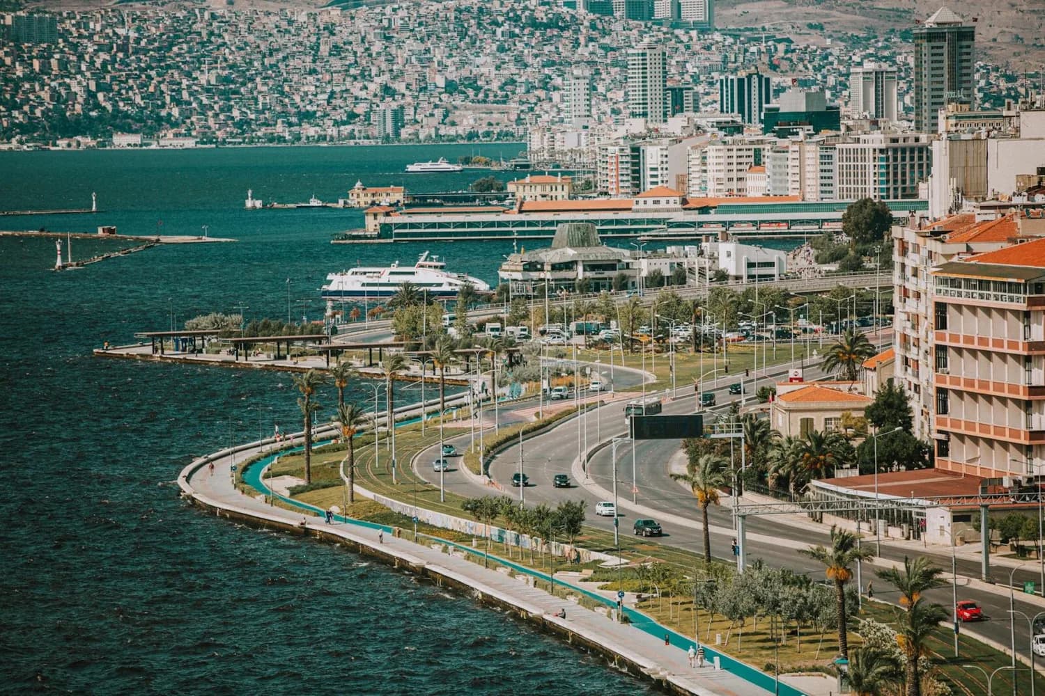 Izmir Küstenpromenade mit Meer, Hafenfähren, Palmen und Uferstraße vor dichter Stadtkulisse