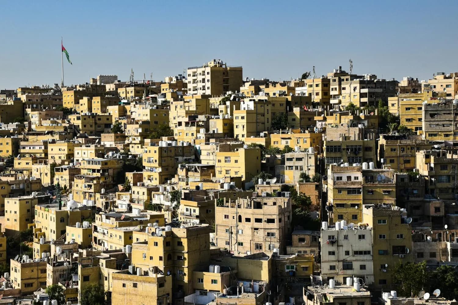 dichtes Wohngebiet in Amman mit gelblichen mehrstöckigen Gebäuden und jordanischer Flagge im Hintergrund, Aufnahme von Royal Jordanian Flug nach München