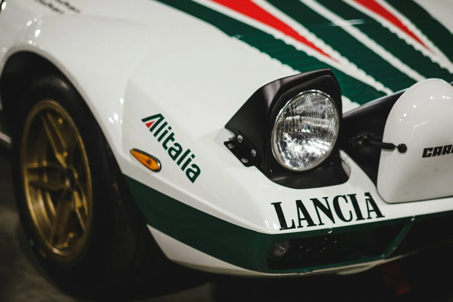 Frontansicht einer weißen Lancia Rallye mit Alitalia Rennlackierung, Scheinwerfer, grünen und roten Streifen sowie goldfarbener Felge