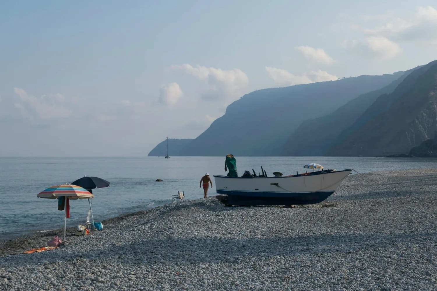 Strand von Reggio Calabria mit Kieselstrand, angerutschtem Fischerboot, Sonnenschirmen und Badegästen vor sanften Küstenklippen und ruhigem Meer (ITA Airways Hinweis)