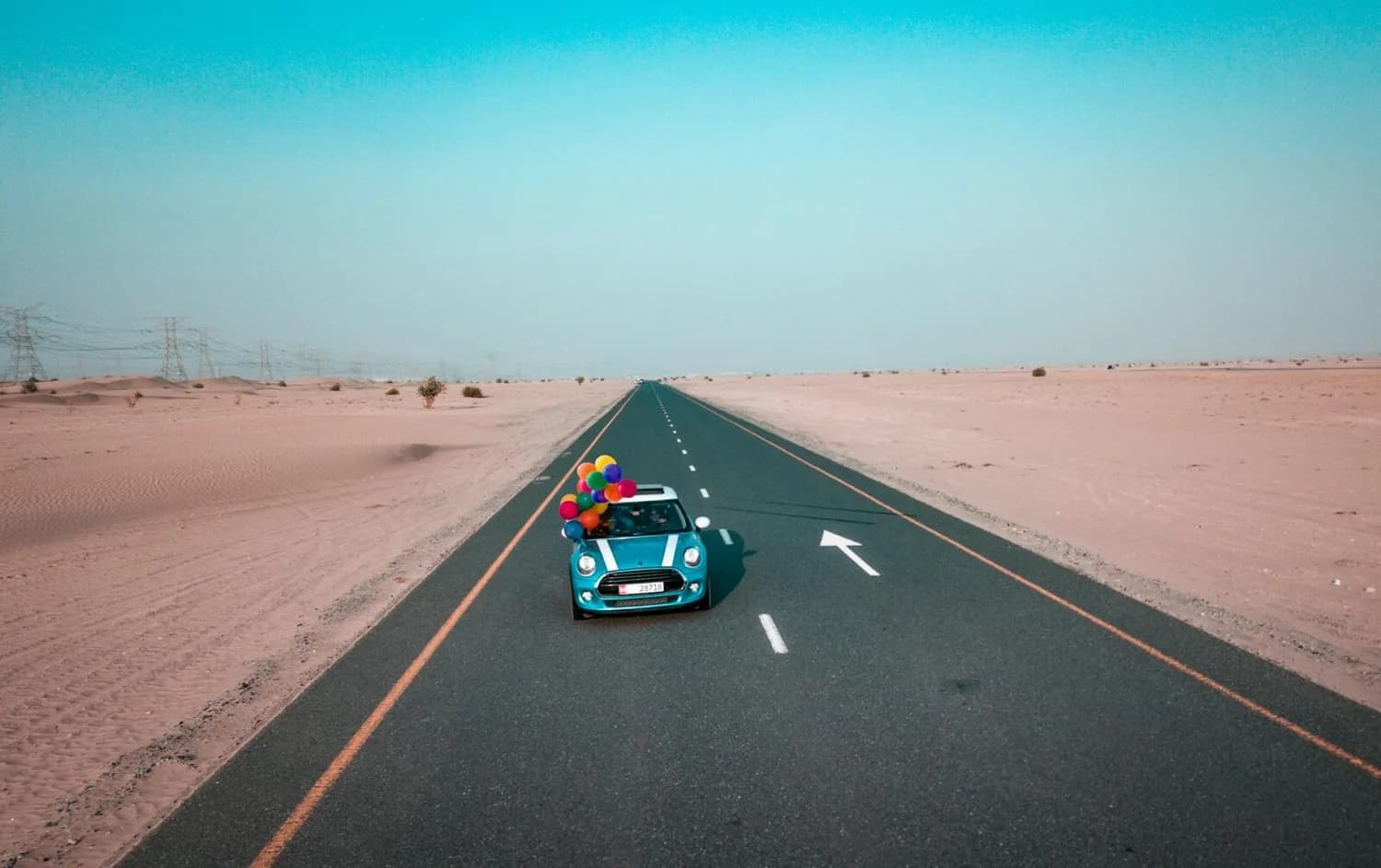 Blaues Mini Cooper Cabrio mit bunten Luftballons fährt allein auf gerader Wüstenautobahn, klarer Himmel, weiter Horizont