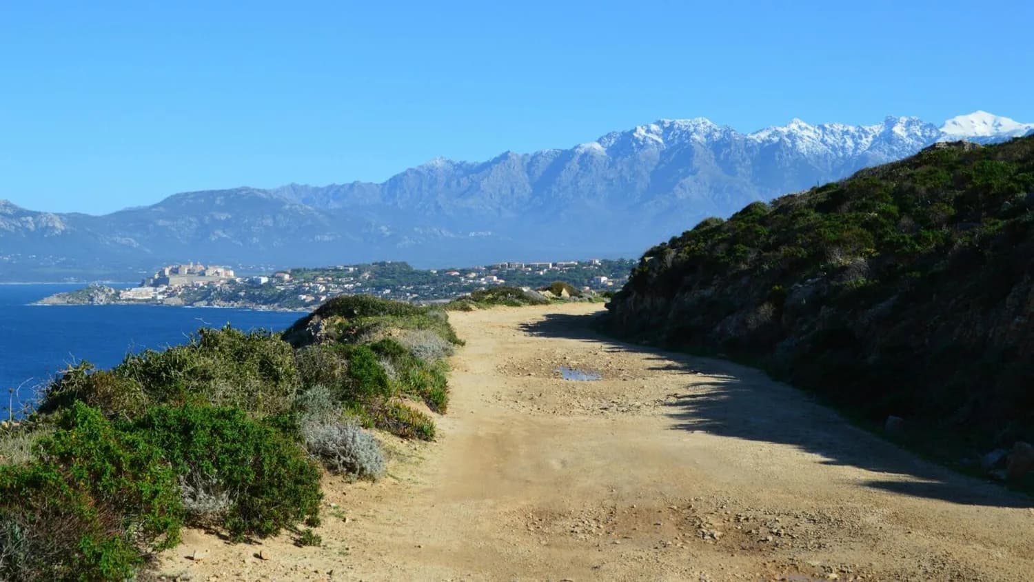 Küstenpfad auf Korsika mit Blick auf das Meer, Küstenort und schneebedeckte Berge im Hintergrund, mediterrane Macchia am Wegesrand