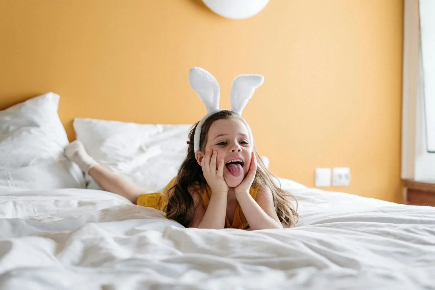 Mädchen mit Osterhasen-Ohren liegt lachend auf Hotelbett in Dortmund während Osterferien