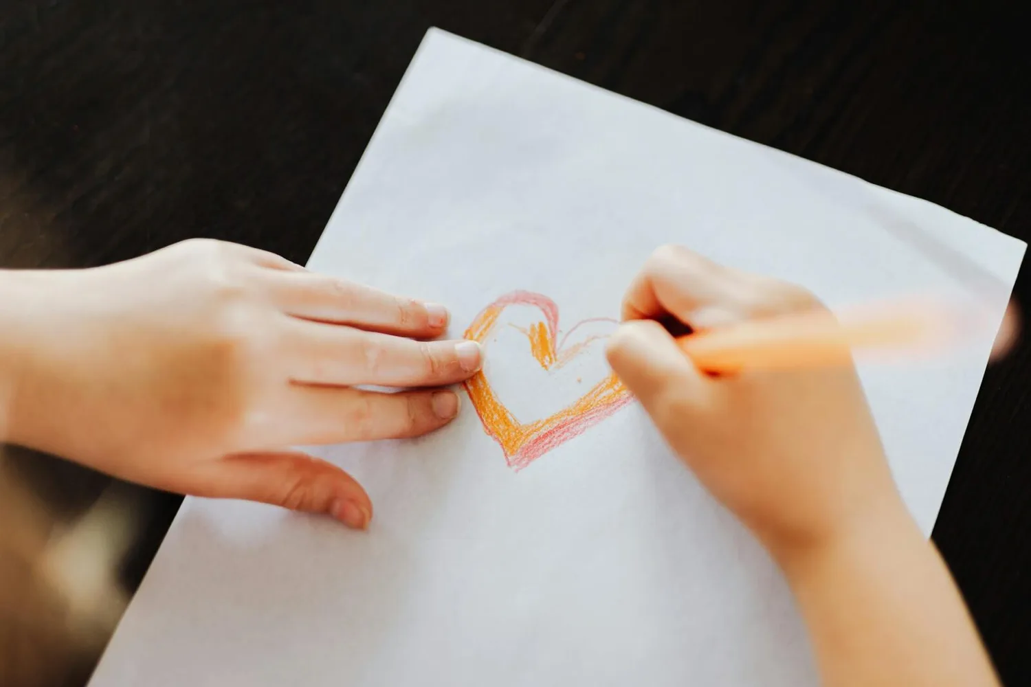 Kinderhände zeichnen buntes Herz mit orangefarbenem Stift auf weißem Papier, EasyJet Easter Collection Produktmotiv, Bastelszene