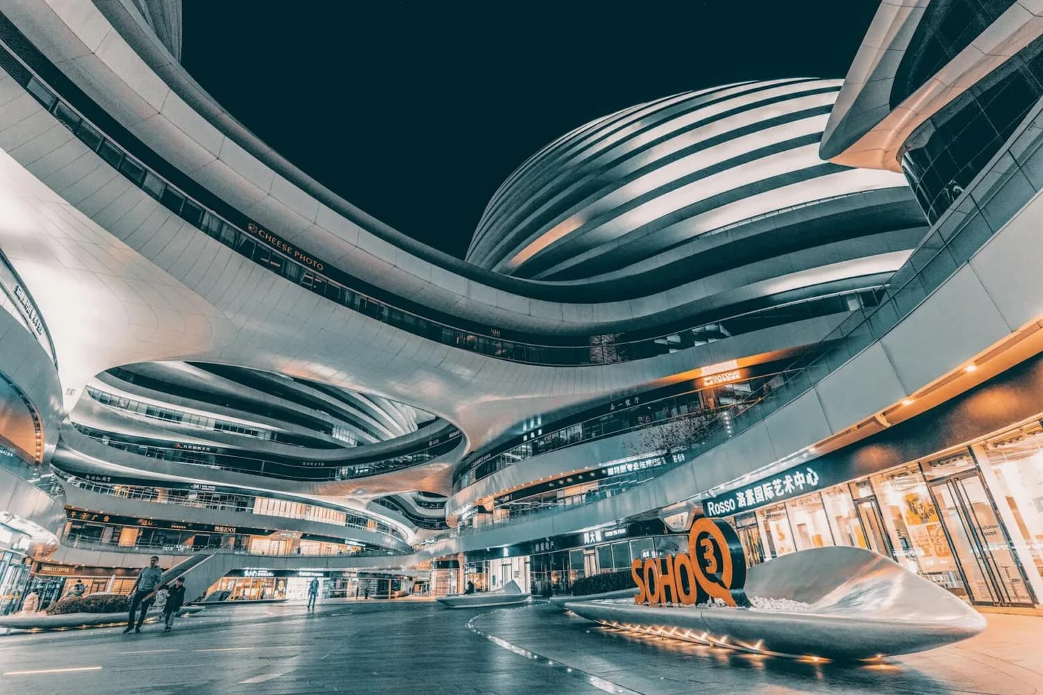 Futuristische geschwungene Fassaden des Galaxy SOHO Einkaufs- und Bürokomplexes in Peking bei Nacht, beleuchtete Ladenfronten und offene Passage mit Skulptur und Personen
