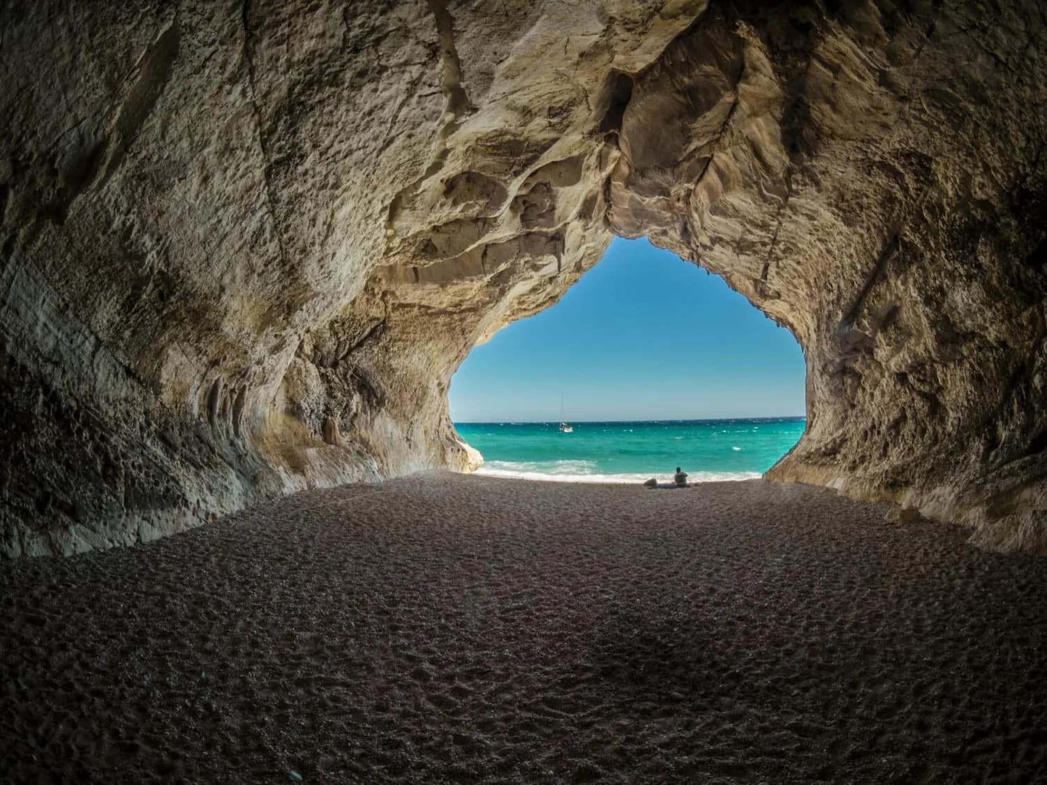 Strandhöhle mit Blick auf türkisblaues Meer, Segelboot am Horizont und Person am Strand – Urlaubsszene Osterferien Flughafen Köln Bonn relevant für Reise und Strandurlaub