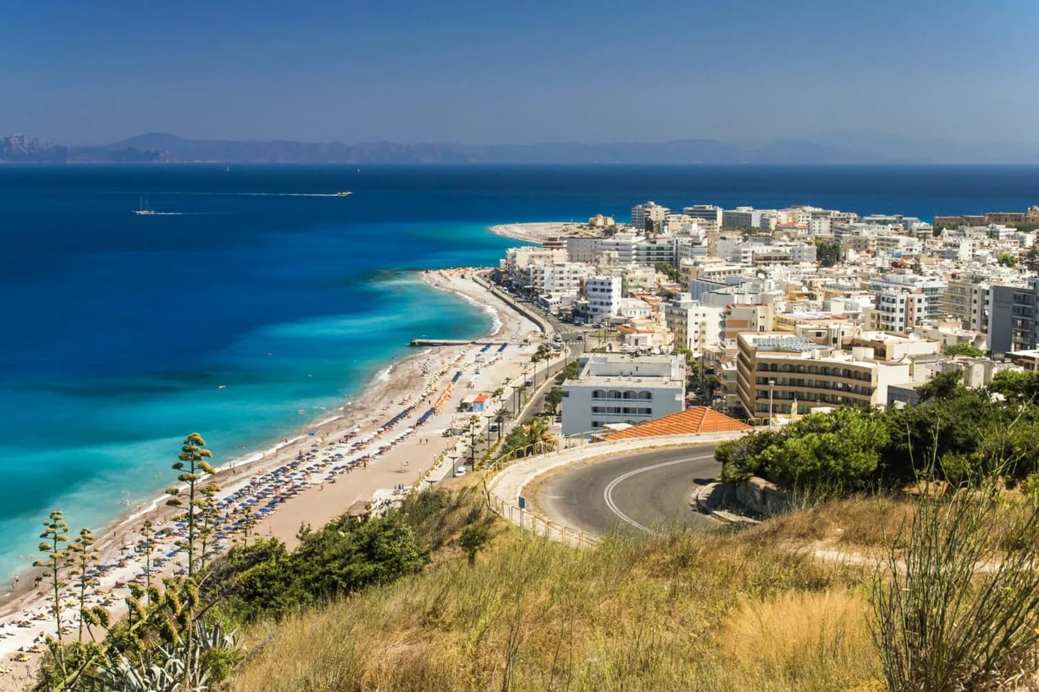 Strandpromenade und Stadtansicht von Rhodos mit blauem Meer, Sandstrand, Sonnenschirmen und kurviger Küstenstraße, Blick aus Richtung München–Rhodos Flugziel technische Details weglassen