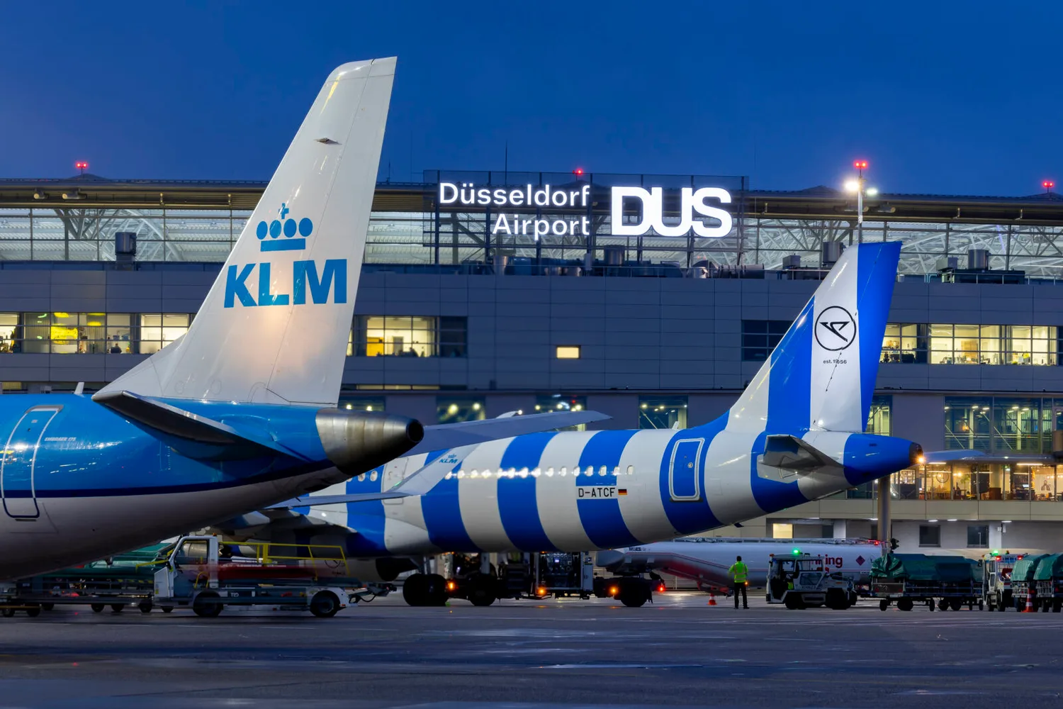 Flugzeughecks mit KLM-Logo und gestreifter Maschine am Nachtpier vor beleuchtetem DUS Düsseldorf Airport Terminal