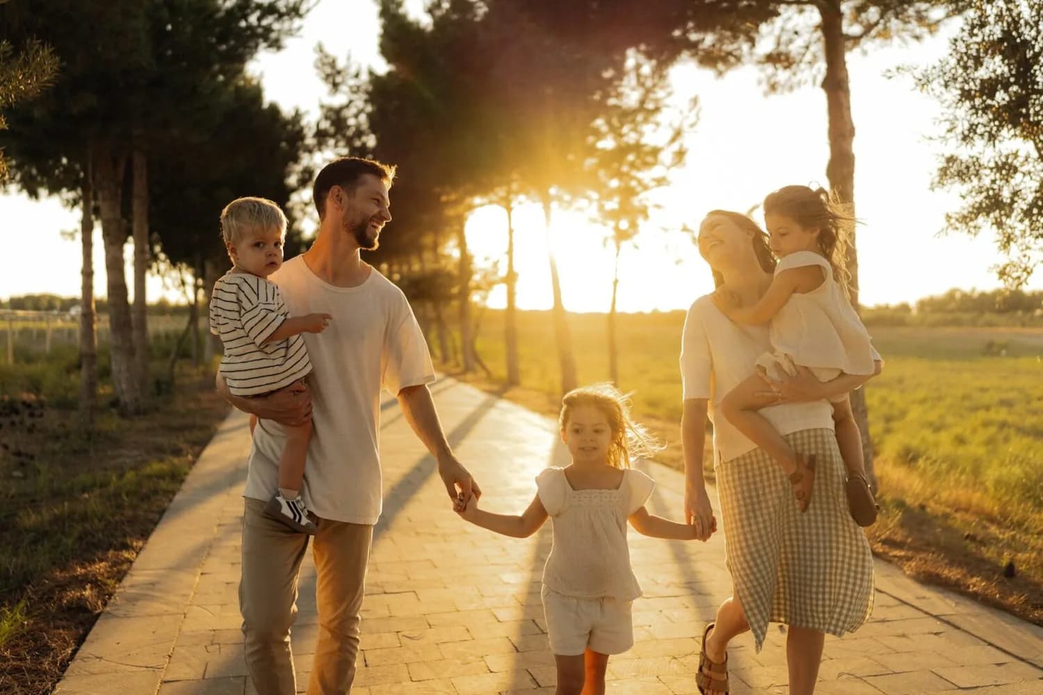Familie beim Spaziergang im Sonnenuntergang, Eltern tragen Kleinkinder und halten die Hand eines Mädchens auf dem Weg durch einen Parklandschaftsweg