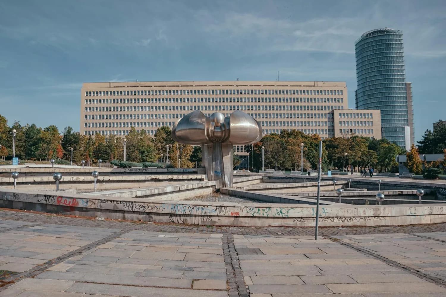 Bratislava Stadtplatz mit modernistischer Metallskulptur in Brunnenanlage vor Verwaltungsgebäude und hohem Büroturm, Fußgänger und Parkumgebung sichtbar