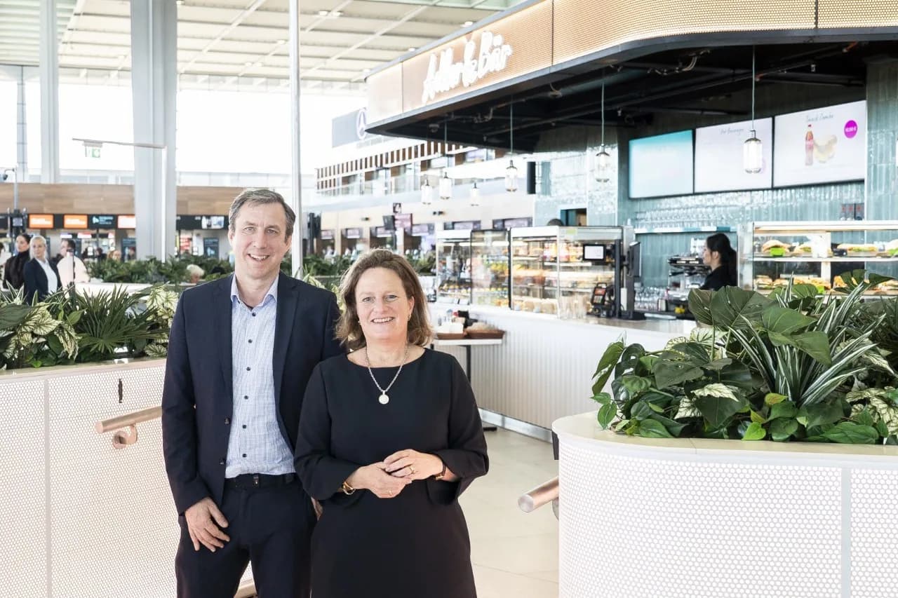 Zwei geschäftlich gekleidete Personen vor Café- und Bistrobereich in moderner Flughafen-Lounge mit Pflanzen und Theke (Adler Bär)