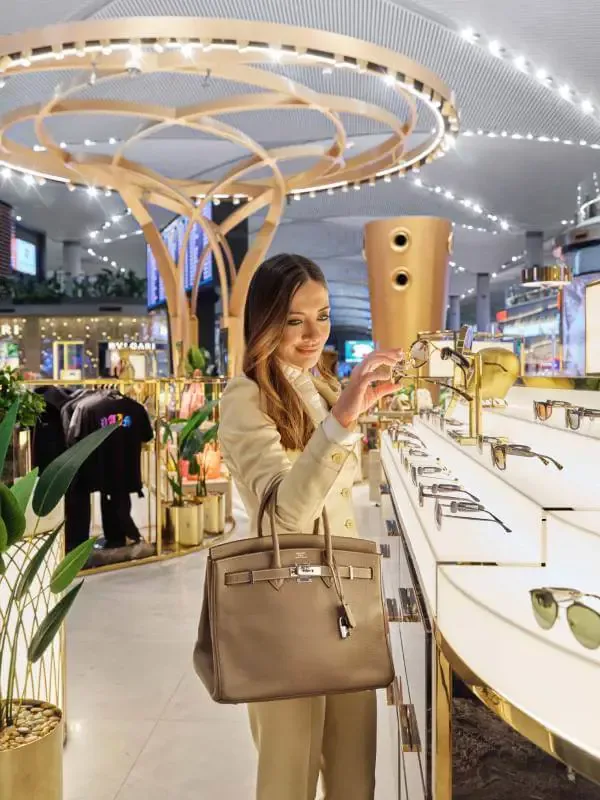 Frau in beigem Mantel shoppt Sonnenbrille an luxuriösem Duty‑Free-Shop-Display im neuem Istanbul Airport Einkaufsbereich, Designer‑Tasche am Arm, moderne Ladenarchitektur im Hintergrund