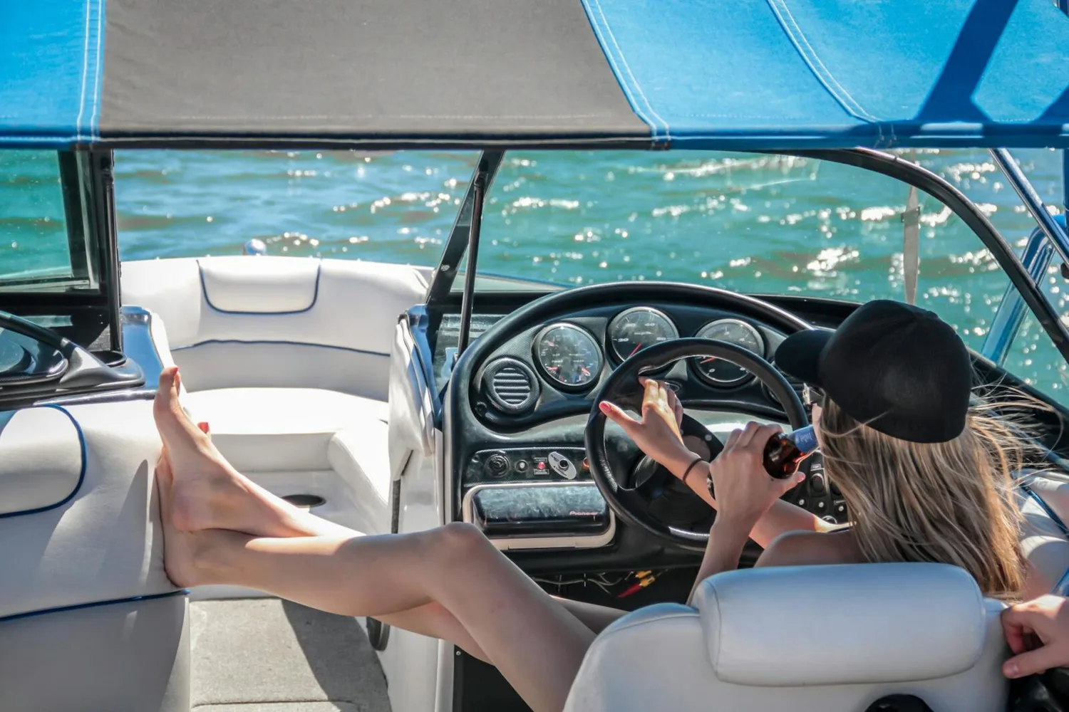 Frau mit Basecap trinkt Bier und steuert entspannt Motorboot auf ruhigem Wasser, Füße auf Polstersitz, Sonnenausblick