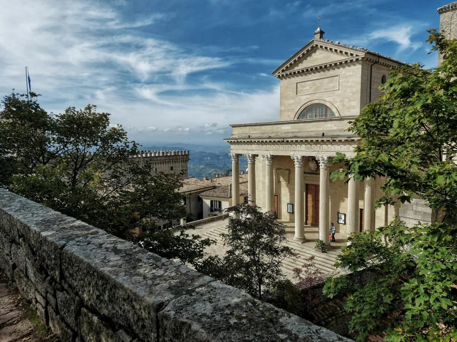 Blick auf die neoklassizistische Basilika und Piazza in San Marino mit Säulenportal, Treppenaufgang, historischen Gebäuden und Hügelpanorama im Hintergrund