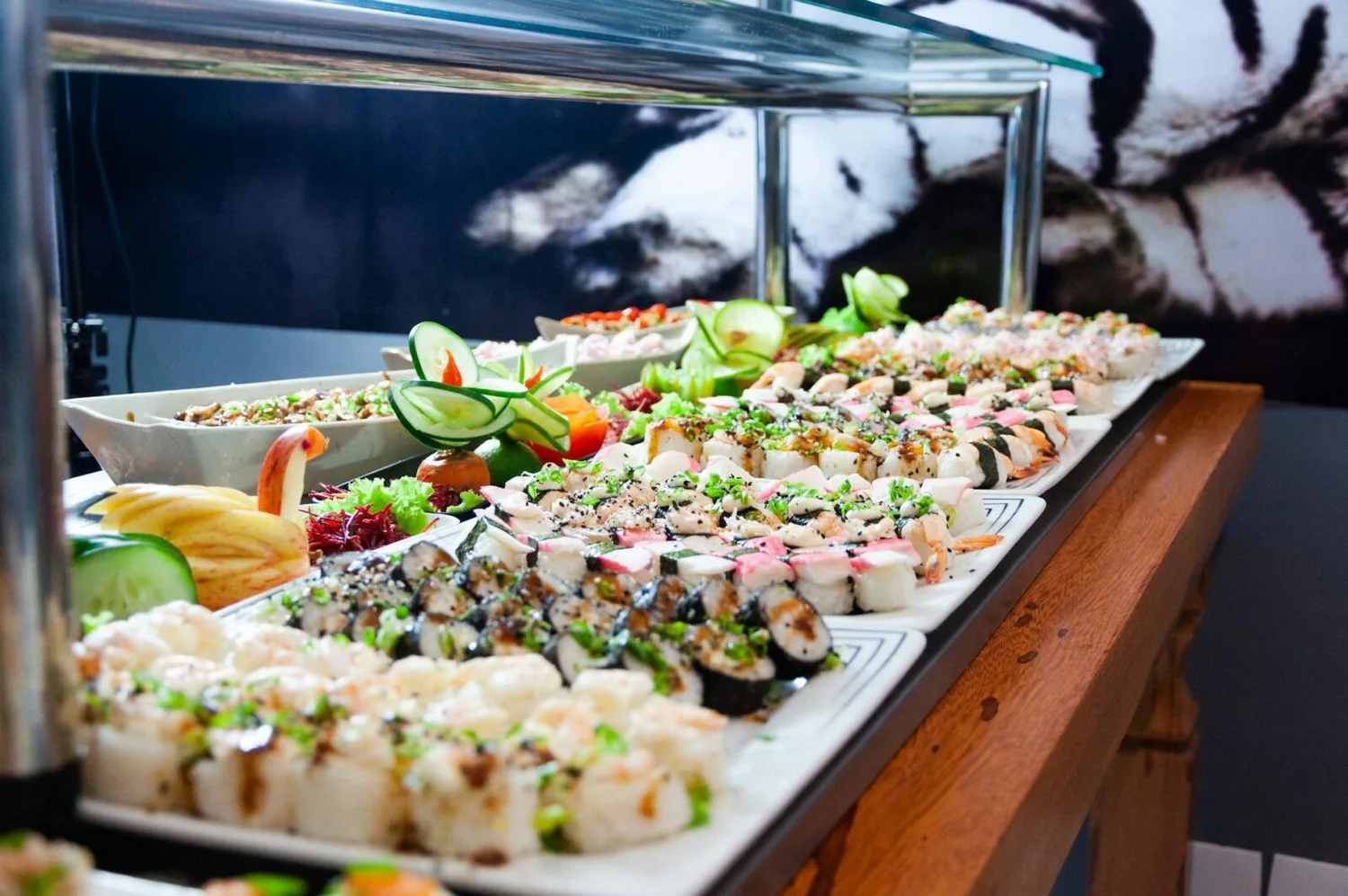 Buffet mit verschiedenen Sushirollen, Garnelen, Kräutern und dekorativen Gurken- und Obstskulpturen auf Buffettisch – Catering Käfer Flughafen BER