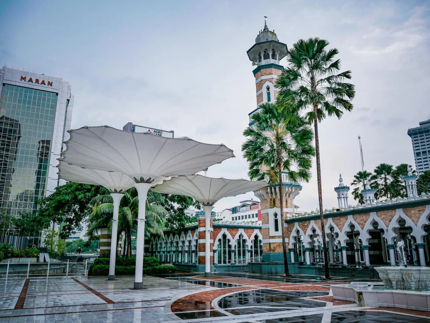 Innenhof der Masjid Jamek in Kuala Lumpur mit Minarett, überdimensionalen Sonnenschirmen, Palmen und umliegenden Hochhäusern
