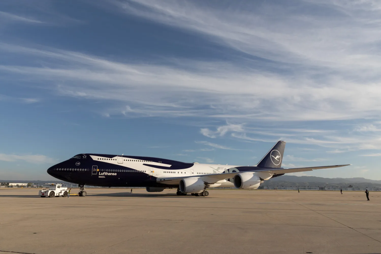 Boeing 747 in Lufthansa XXLkranich Sonderlackierung auf Rollfeld, Seitenansicht mit Schlepper und Flughafenpersonal bei klarem Himmel