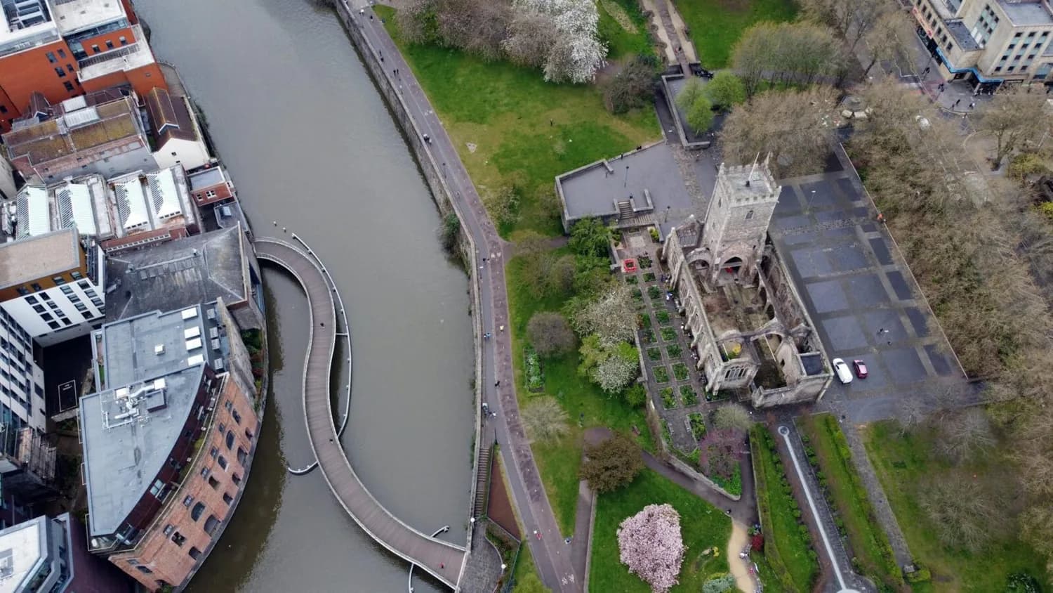 Luftaufnahme eines Flussufers mit geschwungener Fußgängerbrücke, Parkanlage und historischer Kirche, symbolisch für Flugverbindung Köln/Bonn–Bristol