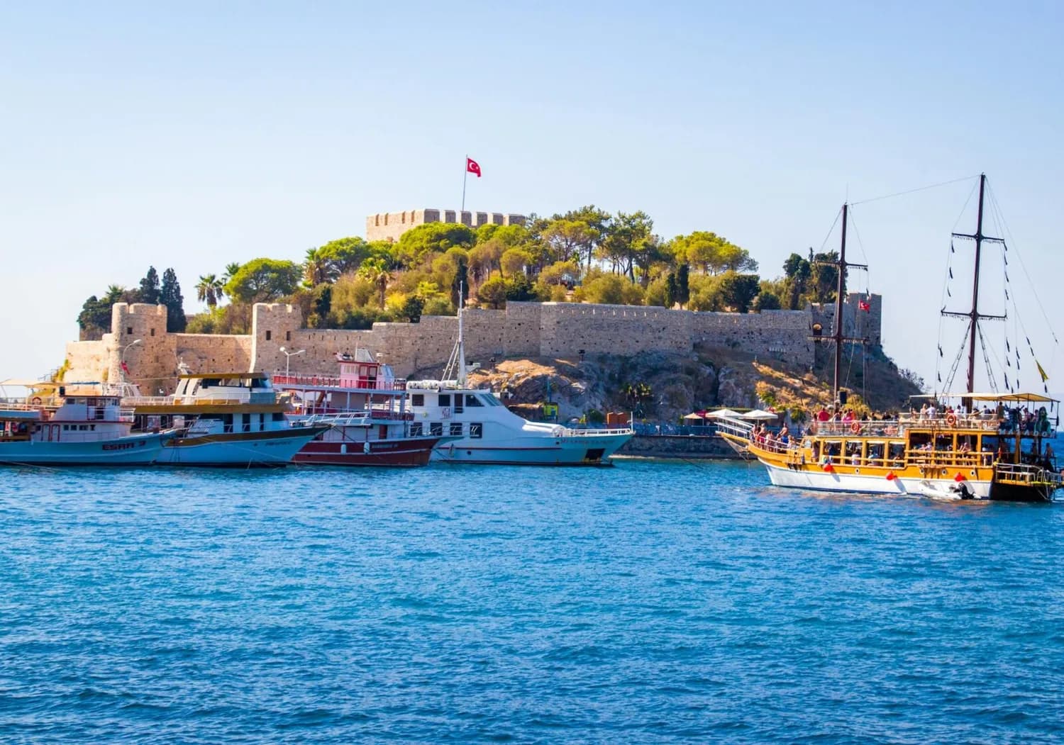 Kuşadası Hafen mit historischen Festungsmauern, türkischer Flagge und Ausflugsbooten auf blauem Meer