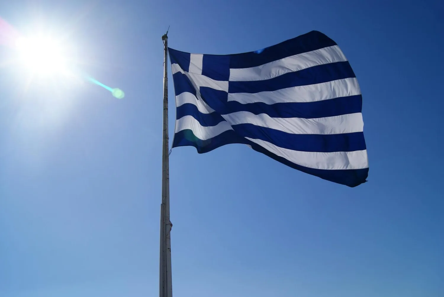 Griechische Flagge mit blau-weißen Streifen weht im Wind vor strahlend blauem Himmel auf Rhodos, Sonne am linken Bildrand