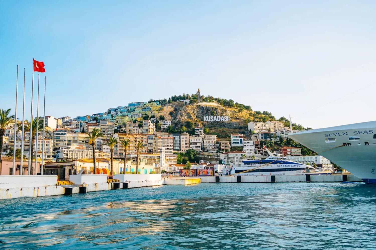 Kuşadası Hafen mit Promenade, ankernden Fähren und Kreuzfahrtschiff vor bunt bebautem Hügel mit Kusadası-Schriftzug und türkischer Flagge