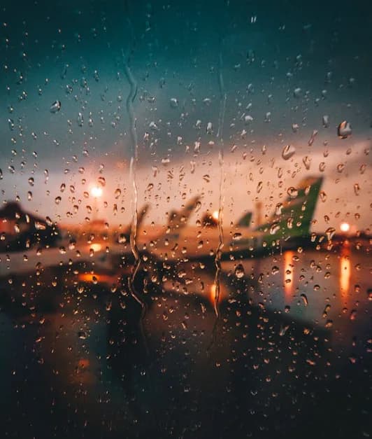 Regenbenetzte Flugzeugfensterscheibe mit Regentropfen, unscharfer Blick auf Flugzeuge und rollendes Gepäck am Flughafen Nürnberg bei Dämmerung, Stimmung von Fernweh und Reisevorfreude