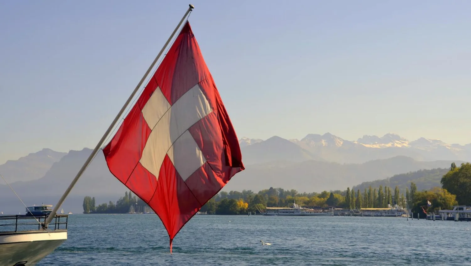 Schweizer Flagge wehend am Seeufer mit Alpenpanorama und Booten, Symbolbild Reform Flughafen Zürich