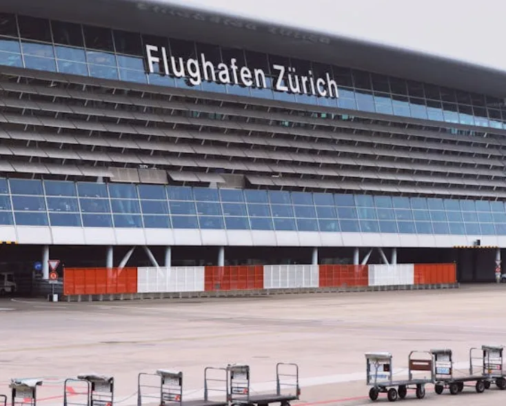 Flughafen Zürich Terminalfront mit großer Beschriftung, Vorfeld mit Gepäckwagen und ParkSleepFly-Bereich im Vordergrund