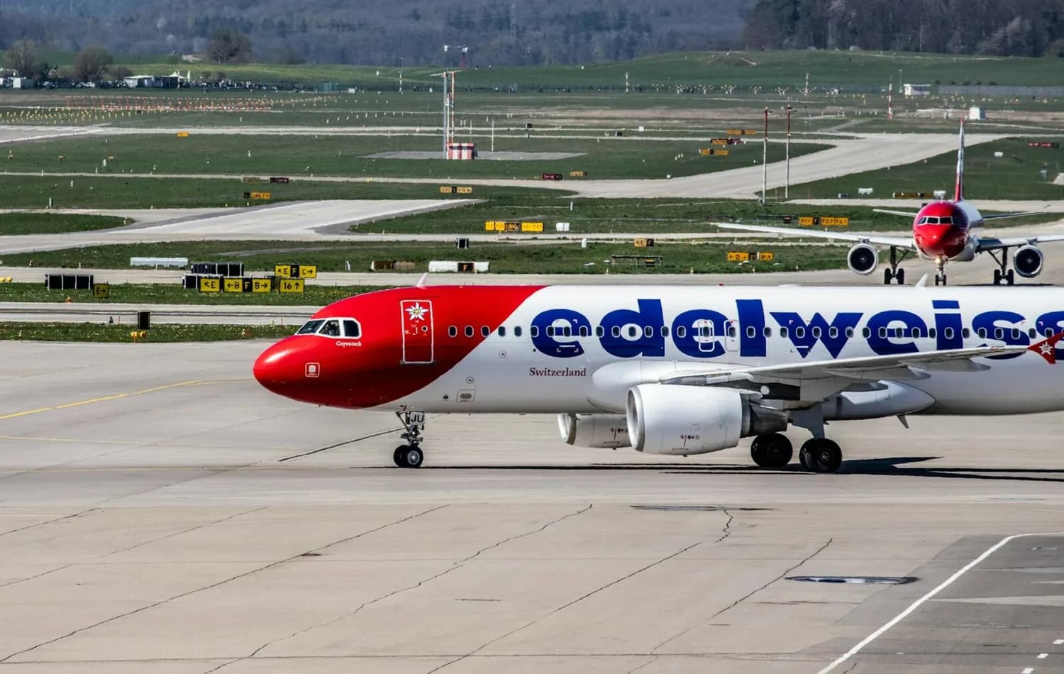 Edelweiss Air Passagierflugzeug mit rot-weißem Seitenrücken und großer Markenbeschriftung auf dem Rollfeld eines Flughafens, im Hintergrund weiteres Taxiing-Flugzeug