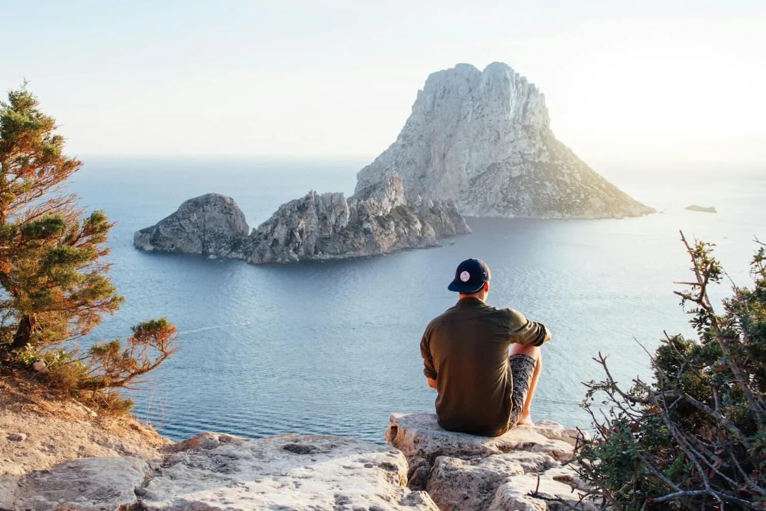 Mann sitzt auf Felskante und blickt auf die Insel Es Vedrà im klaren Mittelmeer, Balearen Last-Minute-Urlaub, Küstenklippen und Sonnenlicht